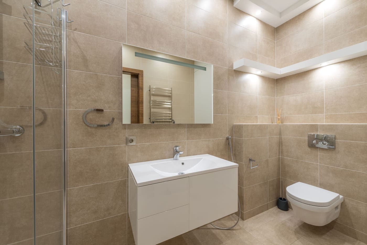 Beige-tiled bathroom lit by recessed ceiling lights and a light strip above the mirror