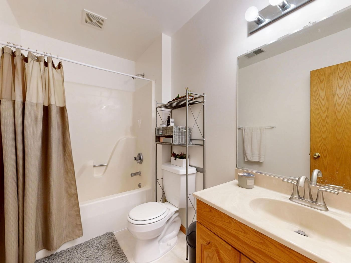 Bathroom with a freestanding metal shelf over the toilet, a beige shower curtain, a gray rug and a vanity