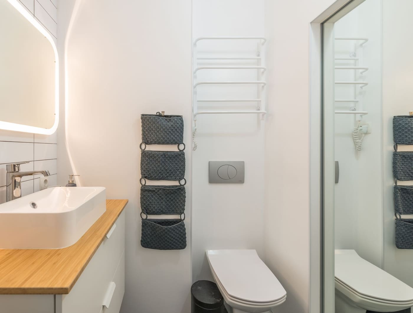 Small white bathroom with a lit mirror, a vessel sink, a hanging fabric pocket organizer and a towel warmer