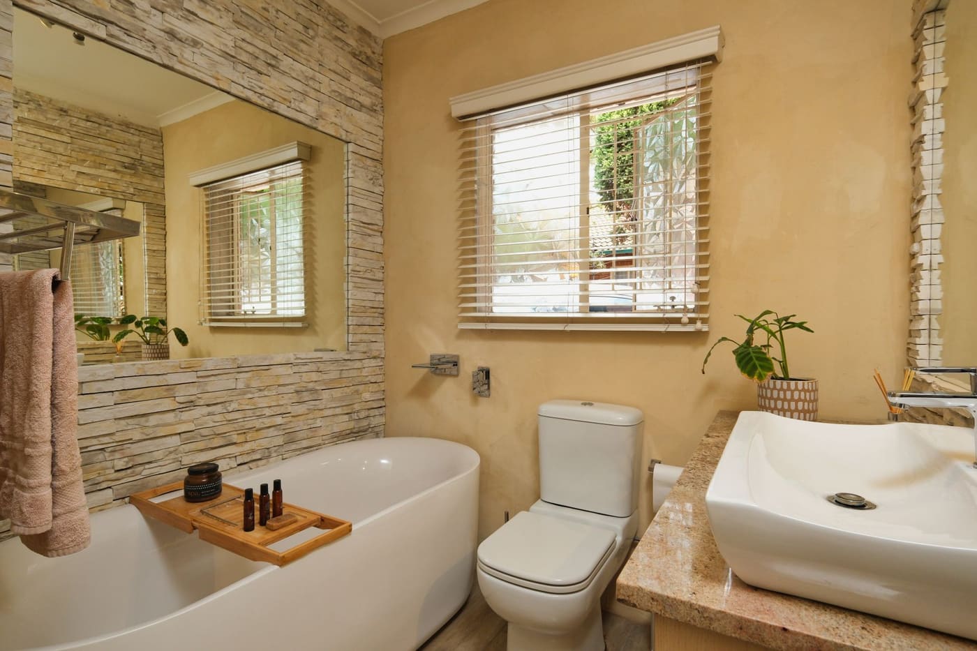 Warm yellow bathroom with a freestanding tub, a wooden tray, a plant and a window